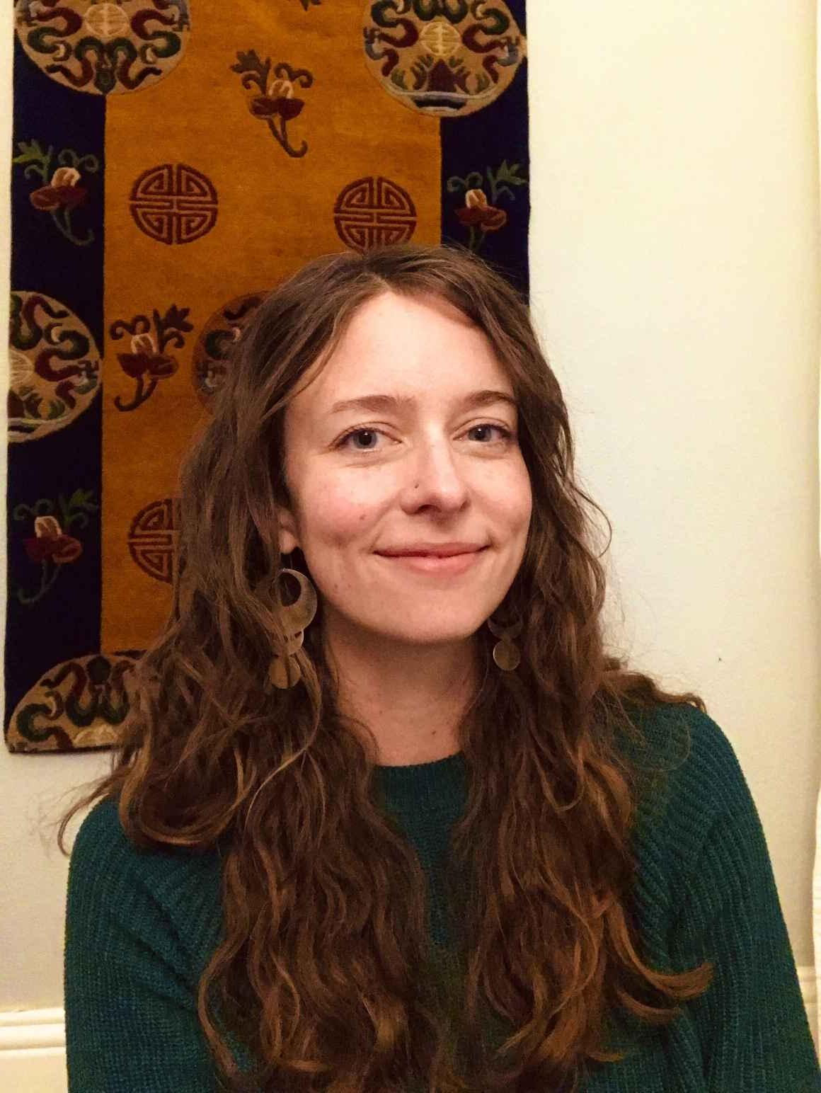 Kate Falkenhart, ACLU of Colorado operations manager, smiling, 在浅色的墙上挂着一幅棕黑相间的挂毯