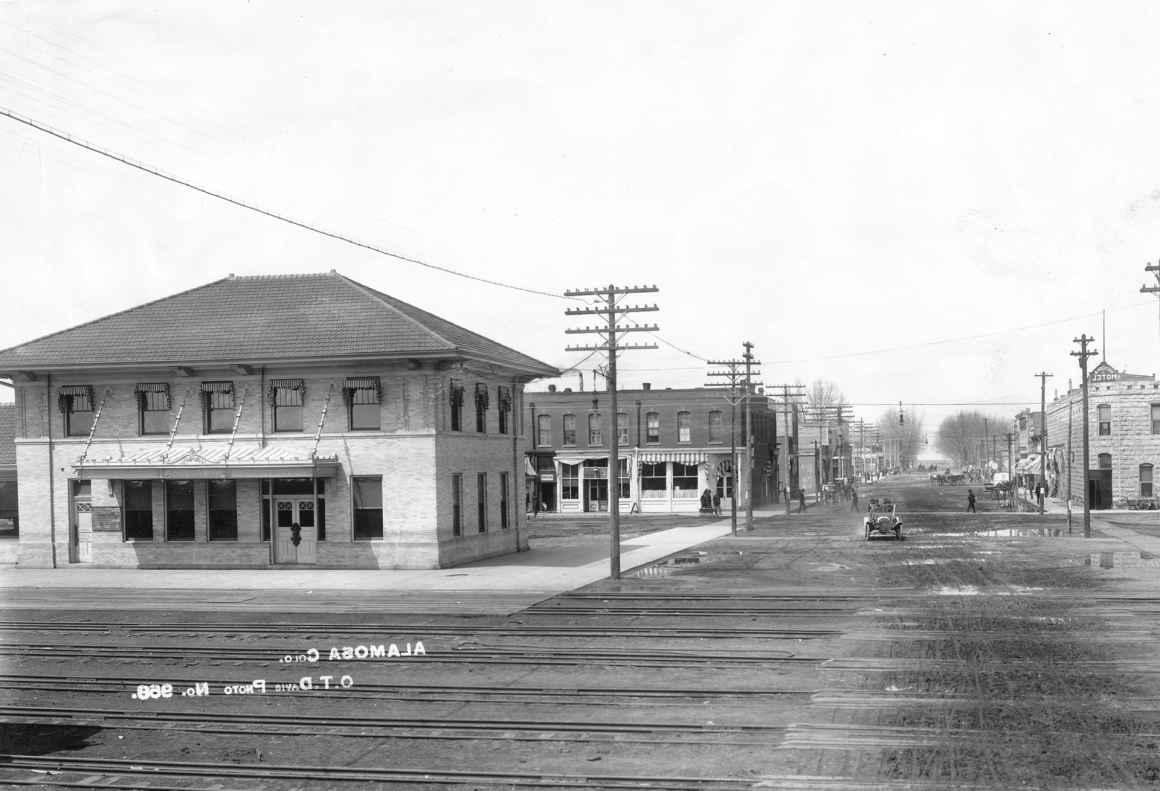 Black and white photograph of a railroad crossing with multiple rails, in a town, next to roads