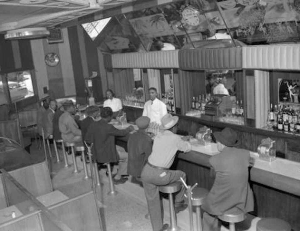 Interior photograph of one male (left) and one female bartending at the 715 Club located at 715 East 26th (twenty-sixth) Avenue in the Five Points neighborhood. A line of male customers fill the seats at the bar. 的y wear fedora and newsboy hats.